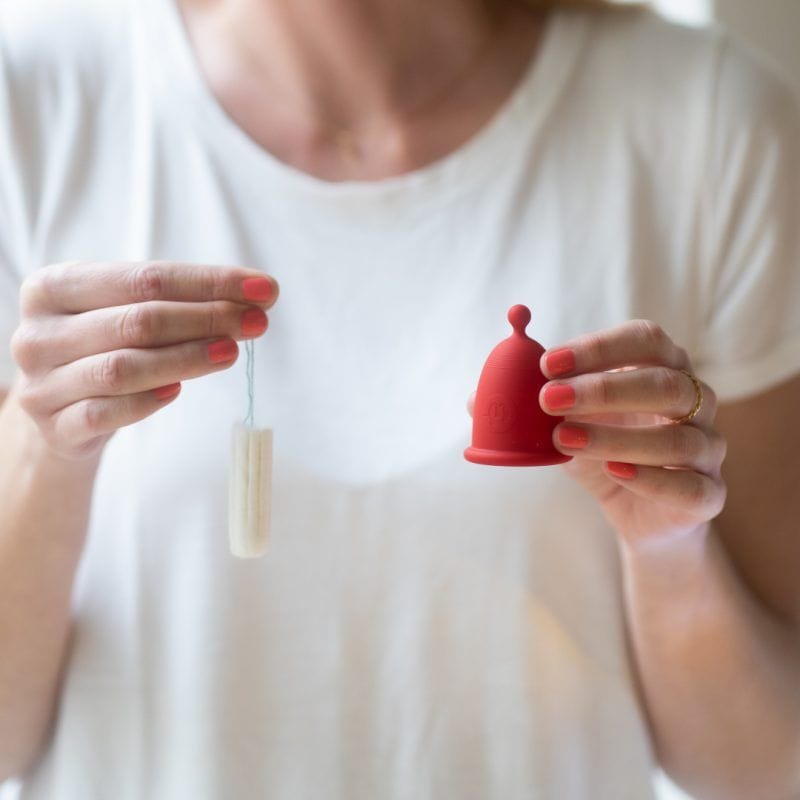 Detailní záběr na ruce ženy, které drží na jedné straně jednorázový tampon a na druhé straně opakovaně použitelný červený silikonový menstruační kalíšek Whoop·de·doo.