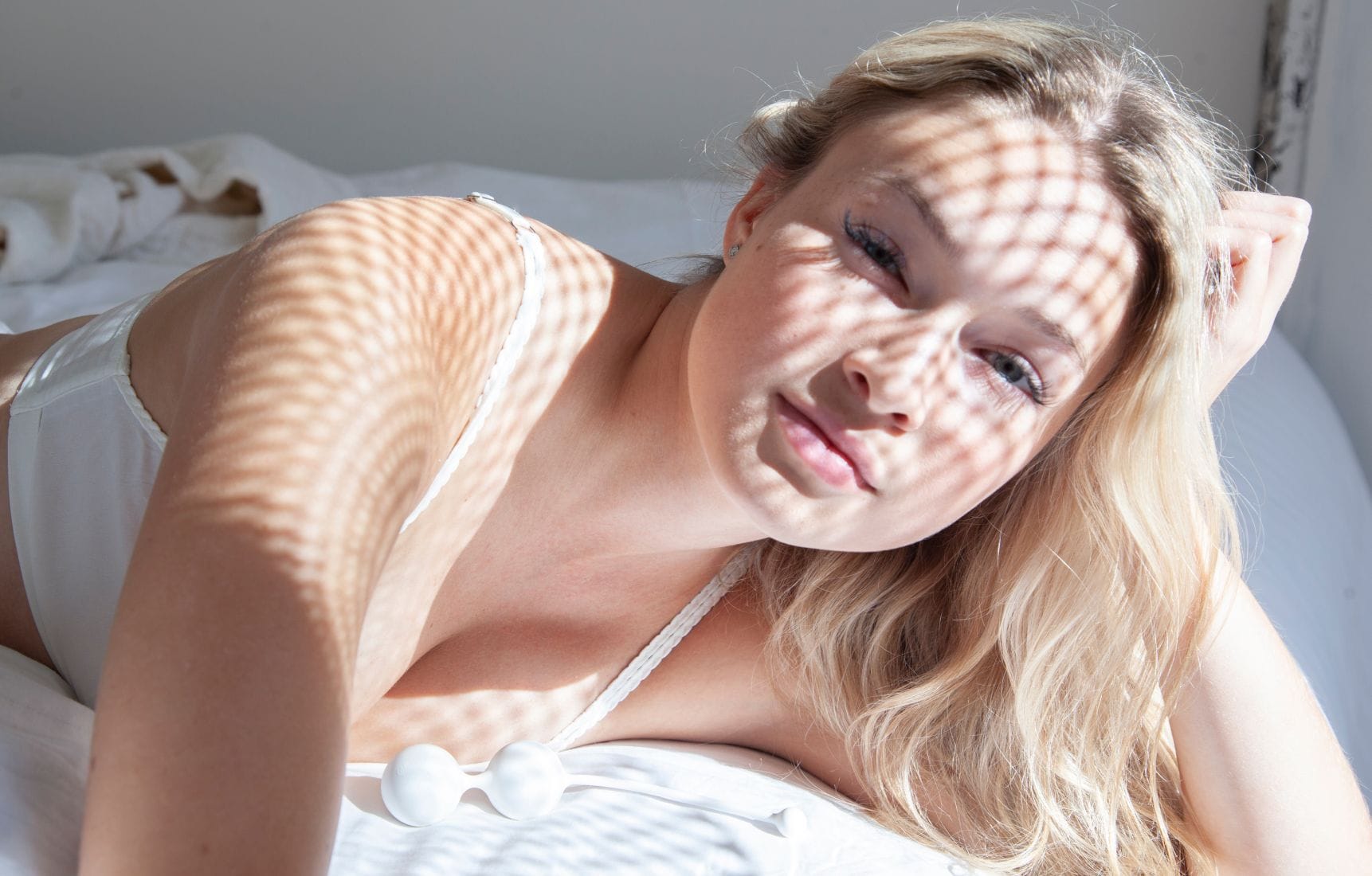 Mladá žena s blond vlasy leží na bílém prostěradle, na kterém jsou položené bílé venušiny kuličky. Přes její tvář dopadá zajímavý stín vytvářející vzor mřížky. Žena působí klidně a uvolněně.
