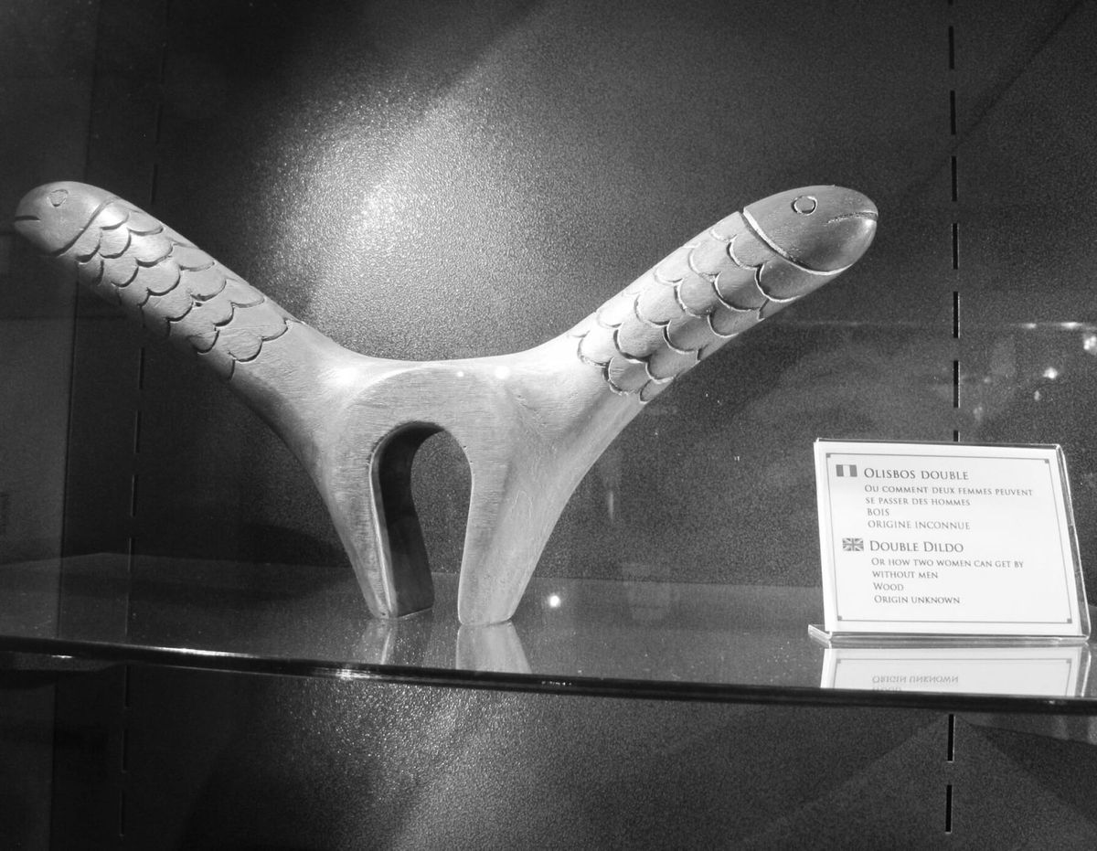 Dřevěný historický dvojitý dildo ve tvaru ryb v muzejní vitríně s popiskem „Olisbos double“.