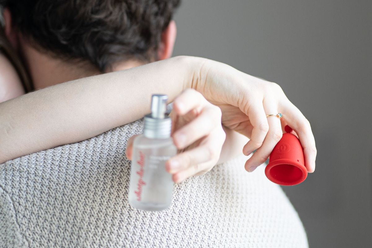Detailní záběr na ruce osoby, která drží lubrikační sprej a červený menstruační kalíšek Whoop·de·doo, zatímco v pozadí objímá druhou osobu.