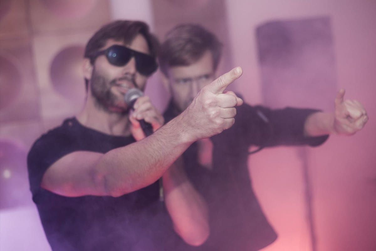 Dva muži vystupující na pódiu, jeden zpívá do mikrofonu v tmavých brýlích a druhý gestikuluje směrem k publiku. Snímek z koncertu kapely 2singles na akci Whoop·de·doo.