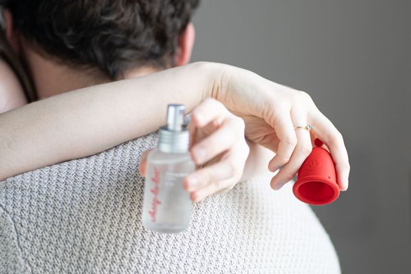 Detailní záběr na ruce osoby, která drží lubrikační sprej a červený menstruační kalíšek Whoop·de·doo, zatímco v pozadí objímá druhou osobu.