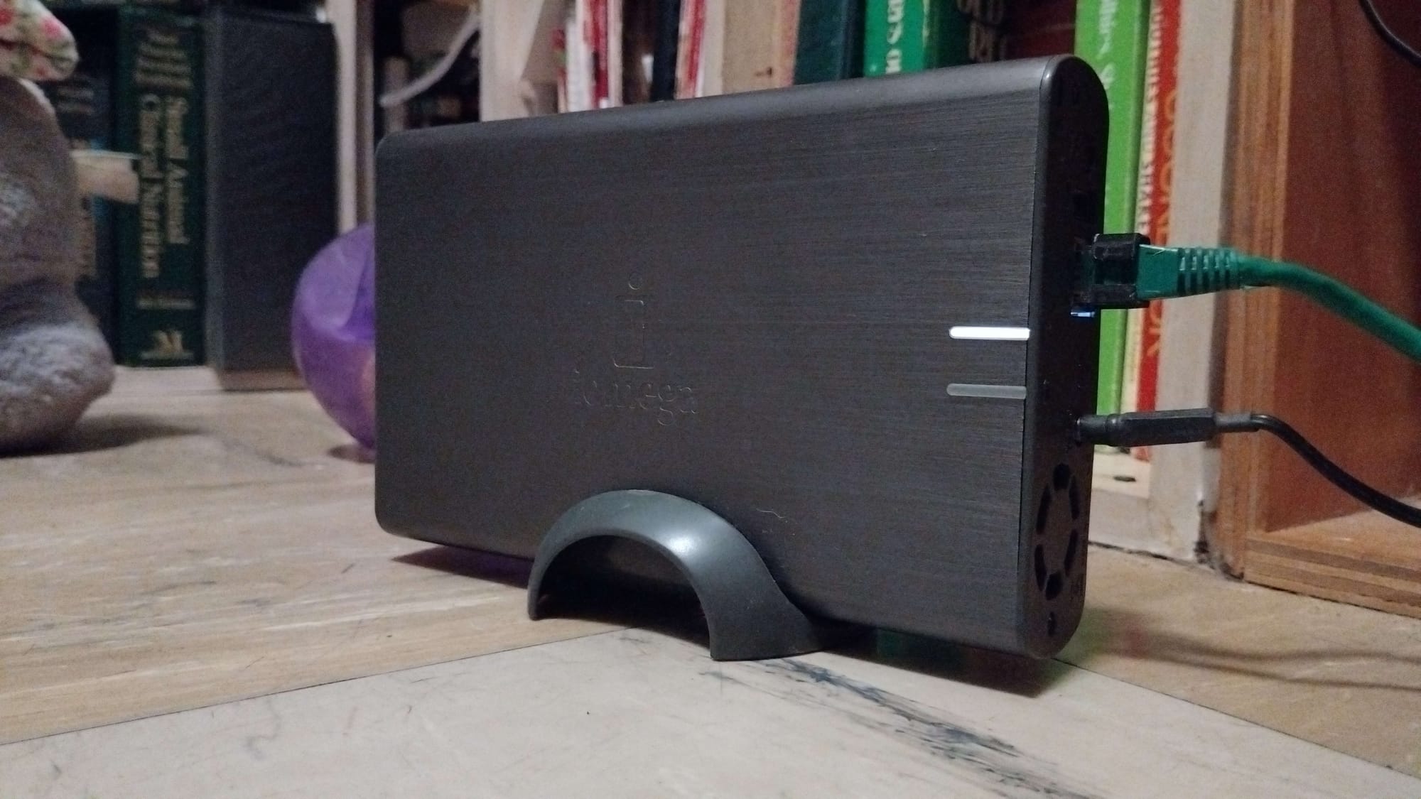 A steel gray color casing that is a NAS. There is a green ethernet cable and a black power cable on the right side, with a white indicator light that shows the status of the NAS itself.