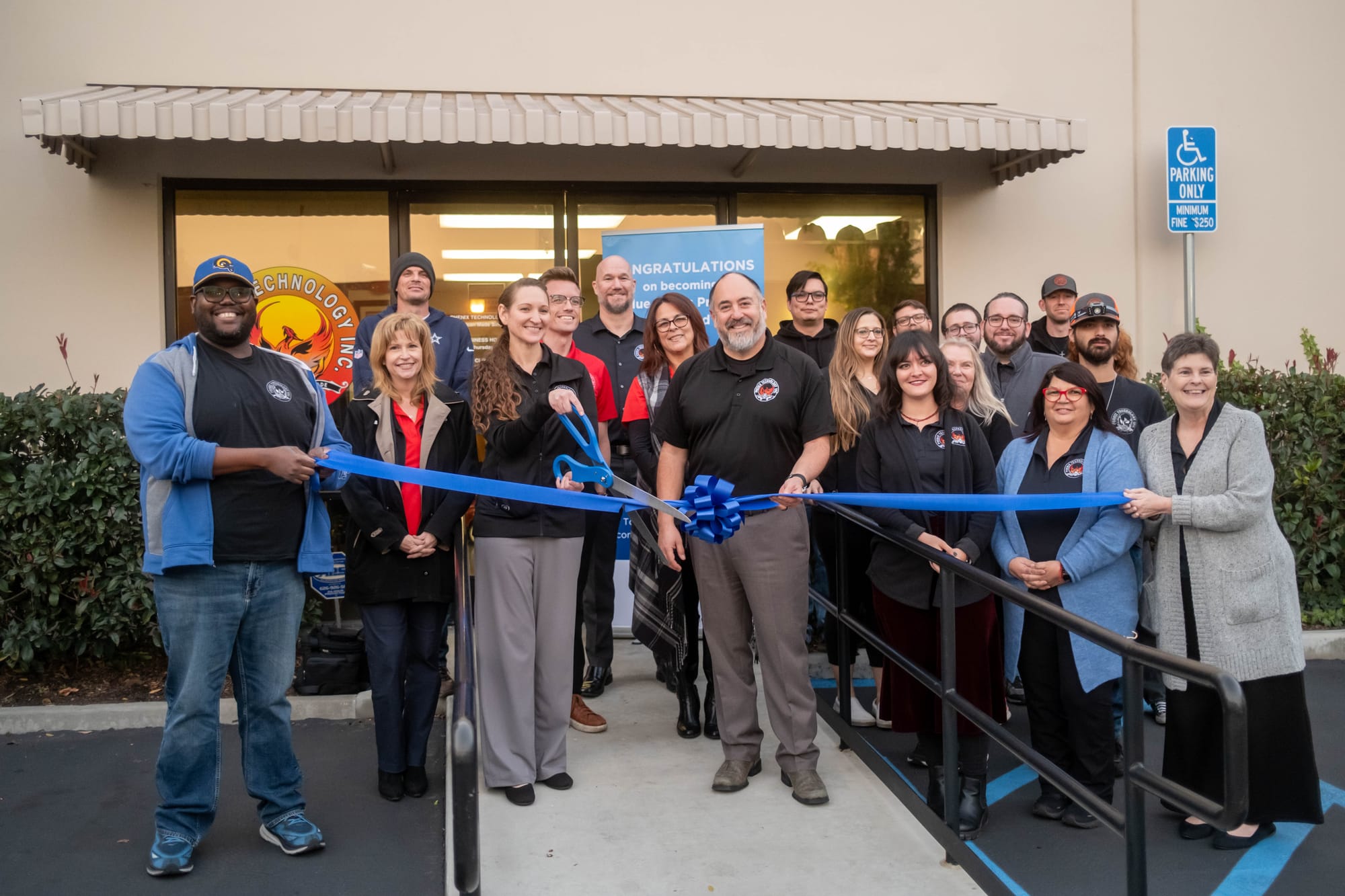 About 20 Phenix Technology employees gather on the front steps of their building for a blue ribbon-cutting ceremony, with a large pair of blue scissors and a congratulatory Blue Zones banner behind them.