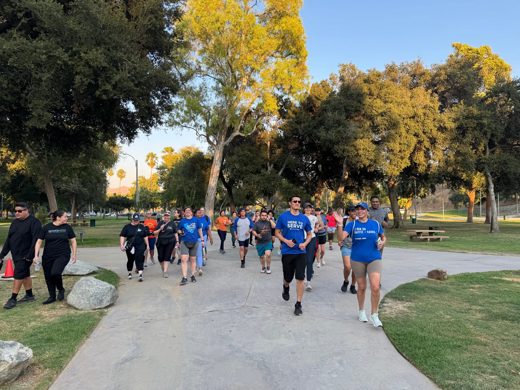 A large group of runners and walkers in blue Blue Zones T-shirts move together along a paved path at Fairmount Park in Riverside on a sunny evening, with mature trees and palm trees in the background.