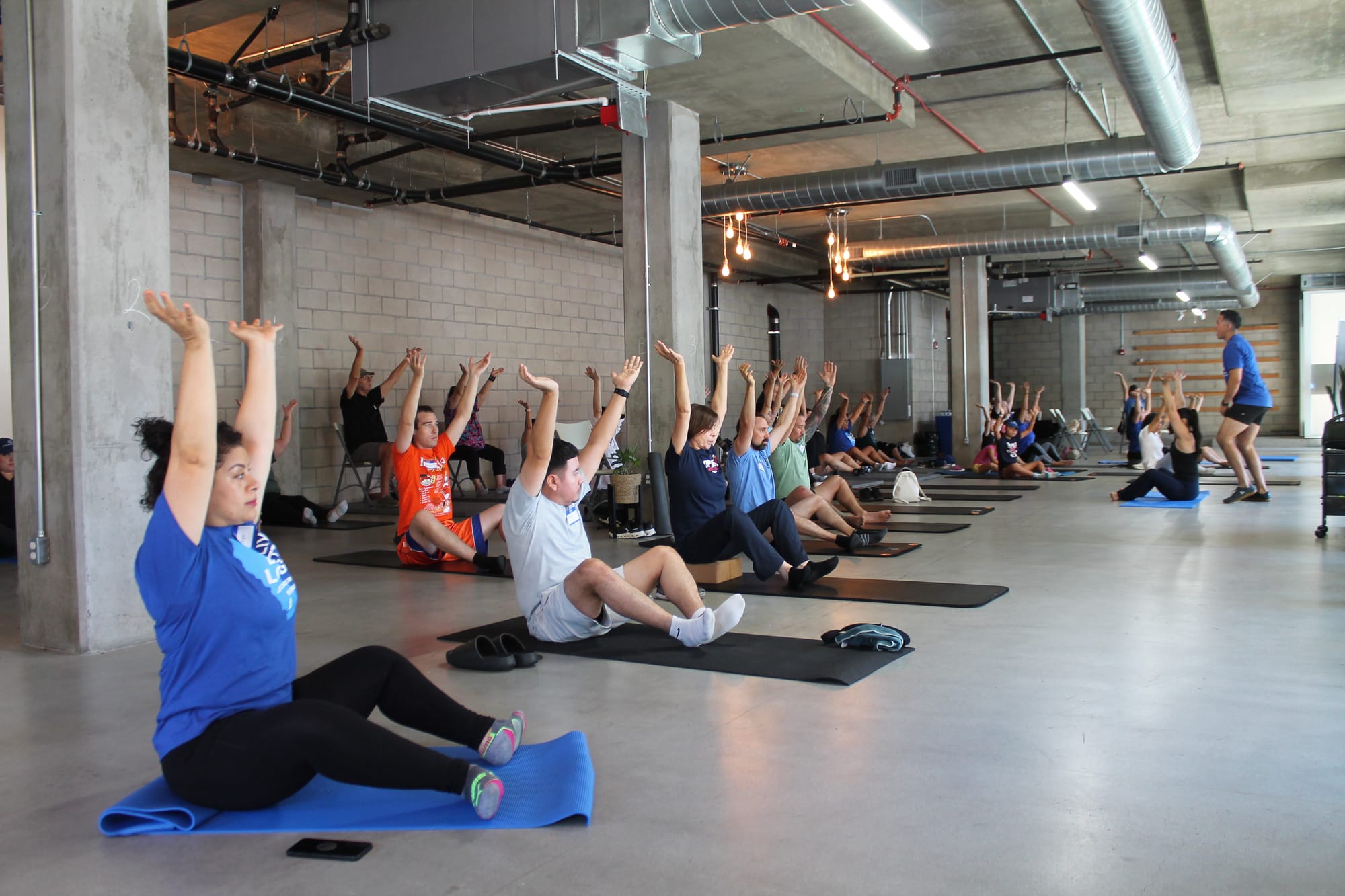 A group of about 20 people sit on yoga mats in a large industrial-style indoor space, stretching with their arms raised overhead, led by an instructor on the right side of the frame.