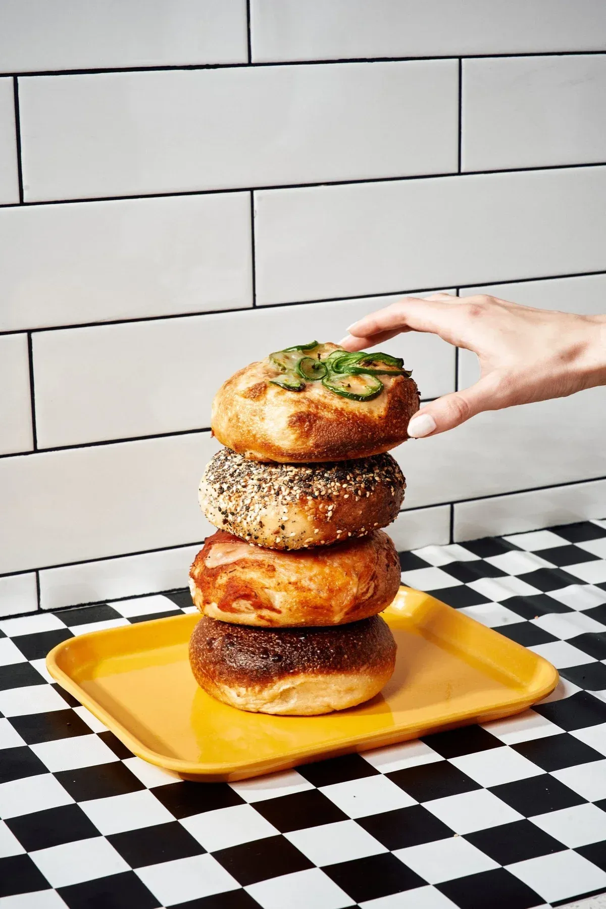 Four house-made New York-style bagels stacked on a yellow tray at Mikey G's in Midtown Riverside: jalapeño, everything, cheese-topped and plain, against white subway tile.