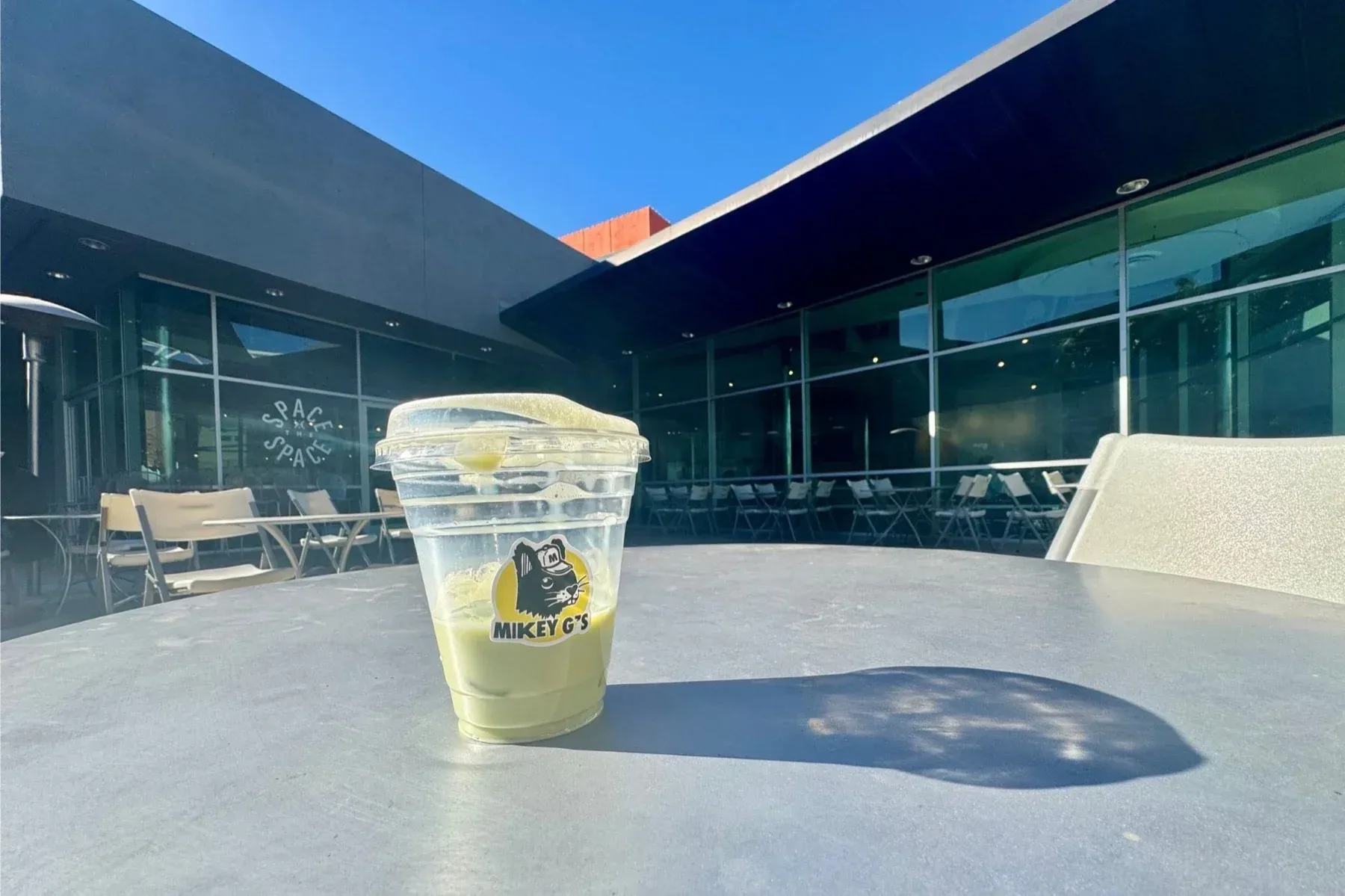 An iced nitro matcha, bright green, in a clear Mikey G's cup printed with the shop's cartoon logo, on a sunlit outdoor patio table in Riverside under a clear blue sky.
