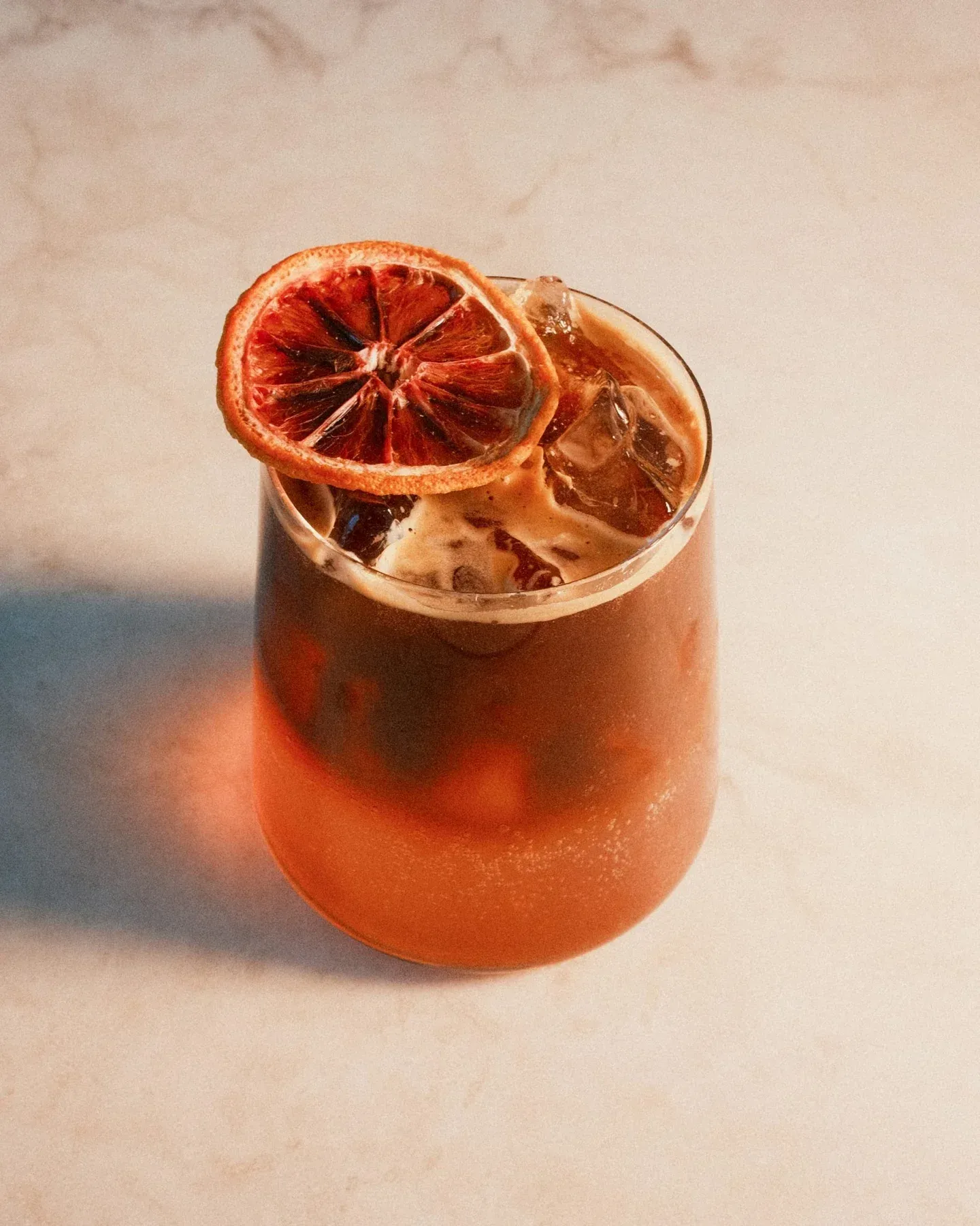 A Blood Orange Espresso Tonic from Mikey G's in Riverside: espresso over tonic water and ice in a stemless glass, dark coffee layered over a glowing orange base, topped with a dried blood orange slice on a pale marble counter in warm light.