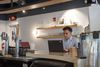 A man works on a laptop at a café counter near drink taps and shelves of glassware.
