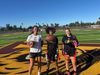 Three girls flag football players stand holding footballs on a field with a tiger mascot logo painted on it.