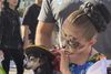 A girl with sugar skull face paint holds a small dog wearing a tiny sombrero hat.