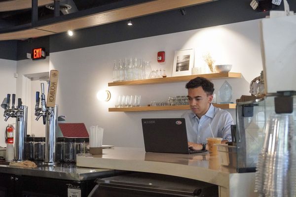 A man works on a laptop at a café counter near drink taps and shelves of glassware.