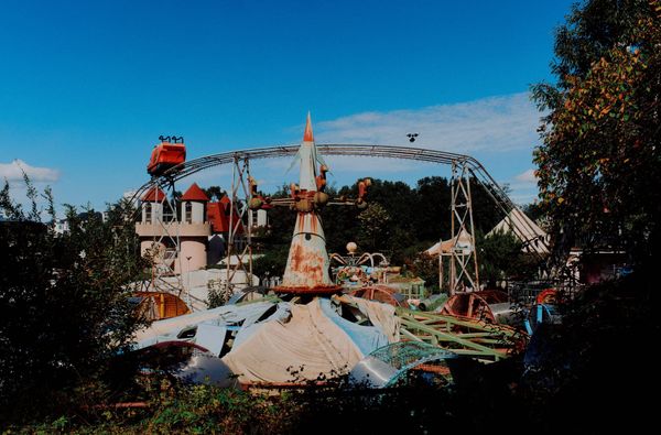 A rusted rocket-shaped ride and overhead monorail track stand amid decaying amusement park rides overtaken by trees.