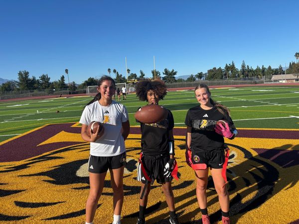 Three girls flag football players stand holding footballs on a field with a tiger mascot logo painted on it.