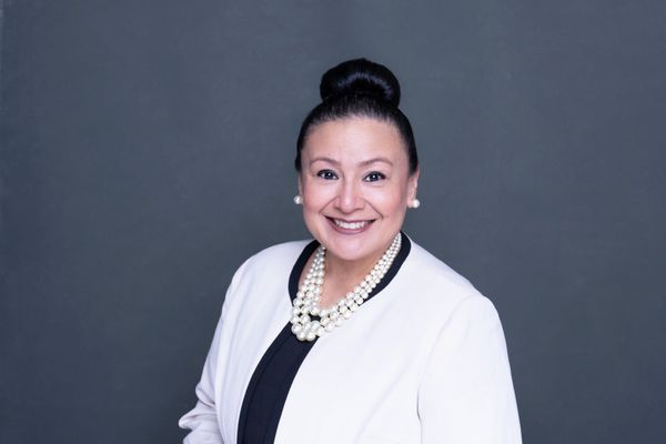 Star Rivera-Lacey smiling in a white jacket with a pearl necklace, against a gray background.