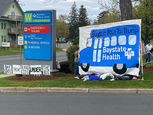 Art work of a large blue bus marked as a Baystate Health bus and the caption "Baystate Bows To Trump". Under the bus canvas are 4 sets of stuffed clothes meant to represent trans children. Se