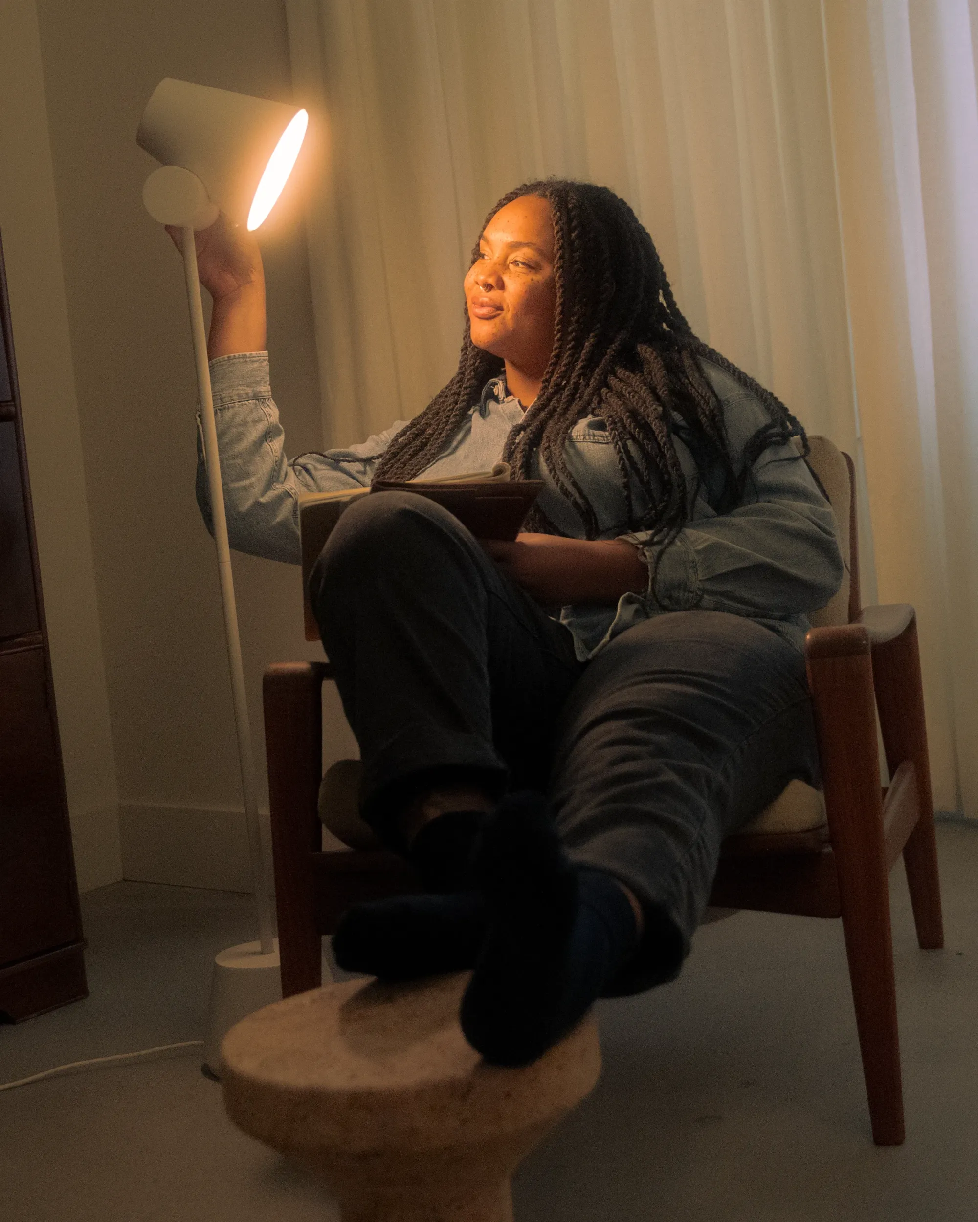 A woman sits on her living room chair. She is holding a tablet and a thin magazine in one hand, and touching Gantri’s Aim Floor Light with the other. The light illuminates her face and frame