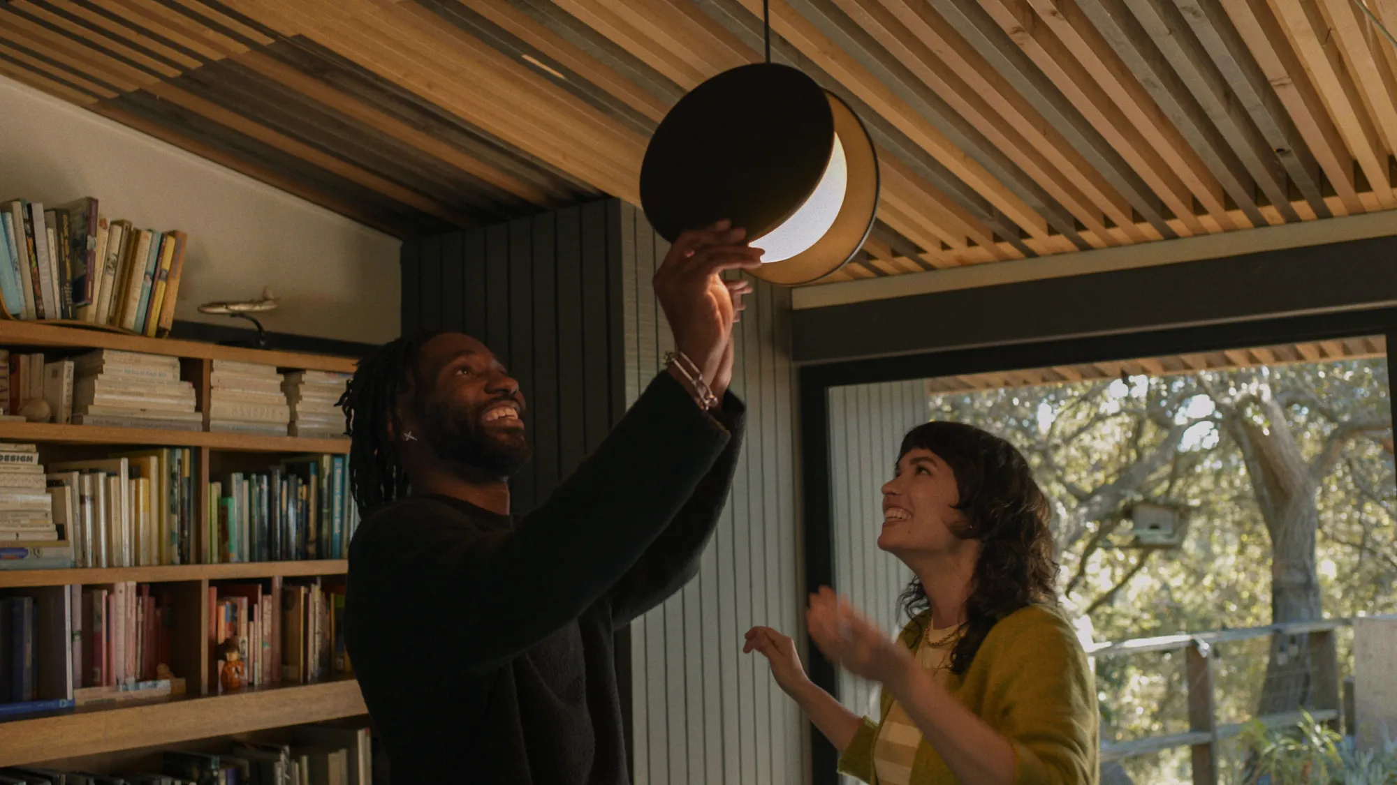 While smiling, a man is fixing his chosen ceiling light with his hands. A woman is also smiling while she looks at their chosen lighting for their space. 