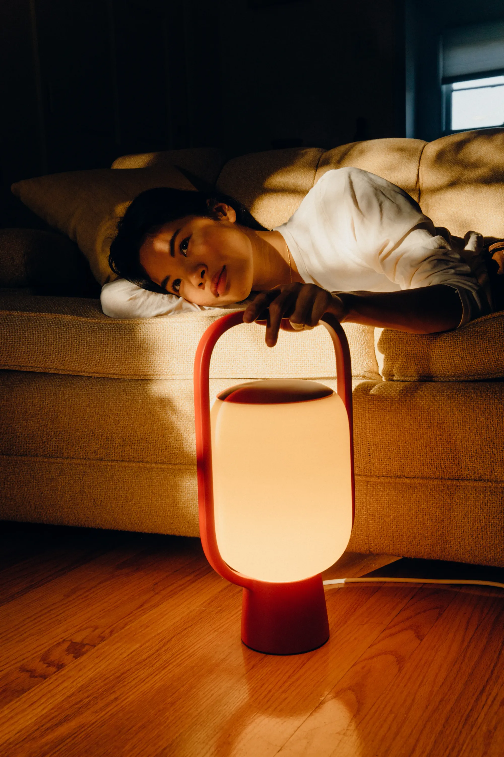 A young woman is smiling slightly while holding on to the top part of a Gantri Kero Table light. Her face is illuminated both by the LED light from the lamp and from a few strays of natural light.