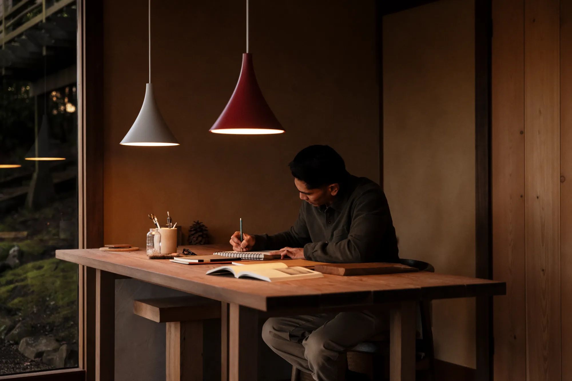 A man is writing intently on a large wooden table. Two ceiling lights are hanging overhead.