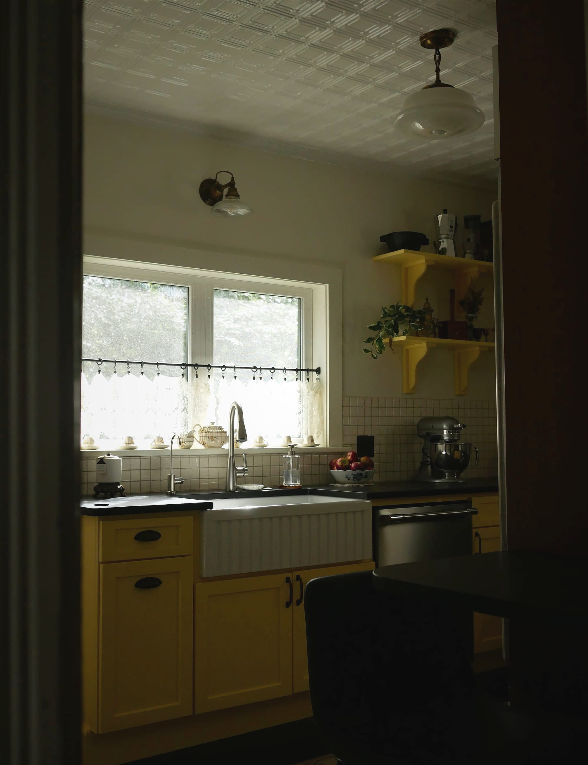 A view of a kitchen sink and window from a doorframe, with yellow lower cabinets in view. 