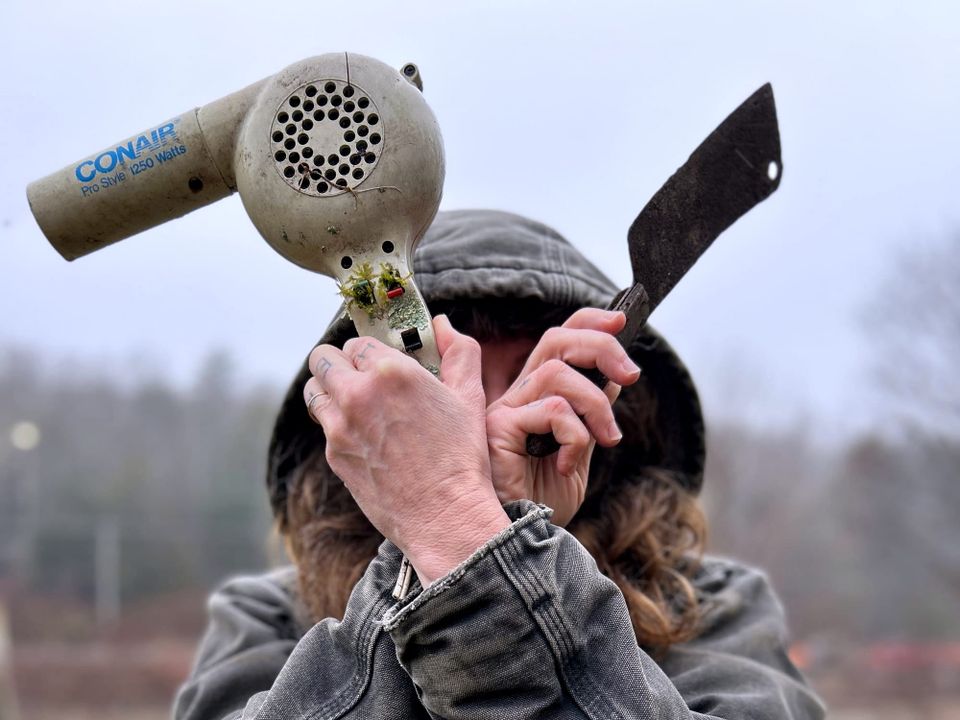 person crossing arms in front of face holding up a Conair hair dryer with moss growing out of the buttons and a rusty meat cleaver, in front of overcast sky