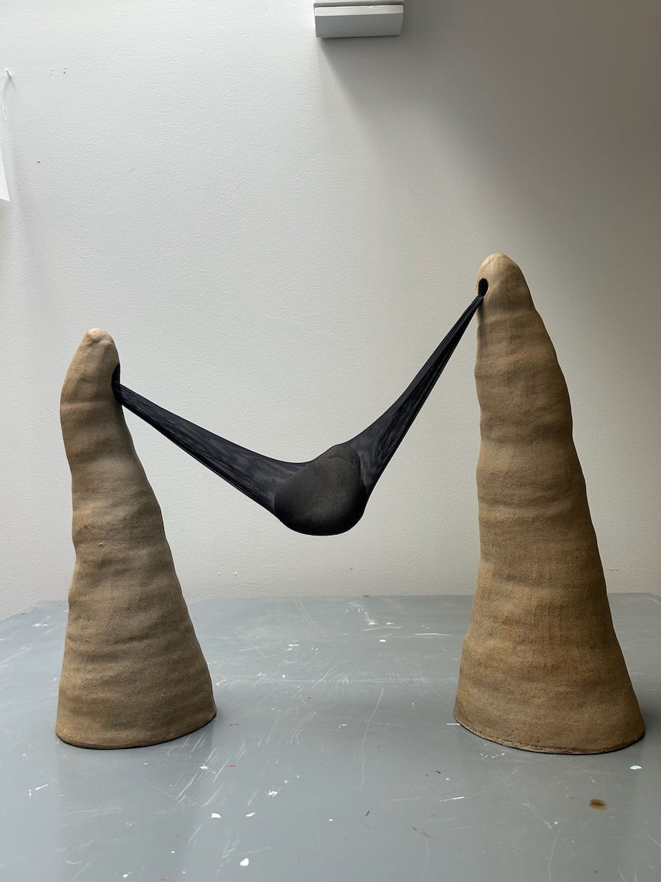 an abstract sculpture made of two lumpy clay cones, with nylon strung between them and a rock inside the nylon so it hangs down