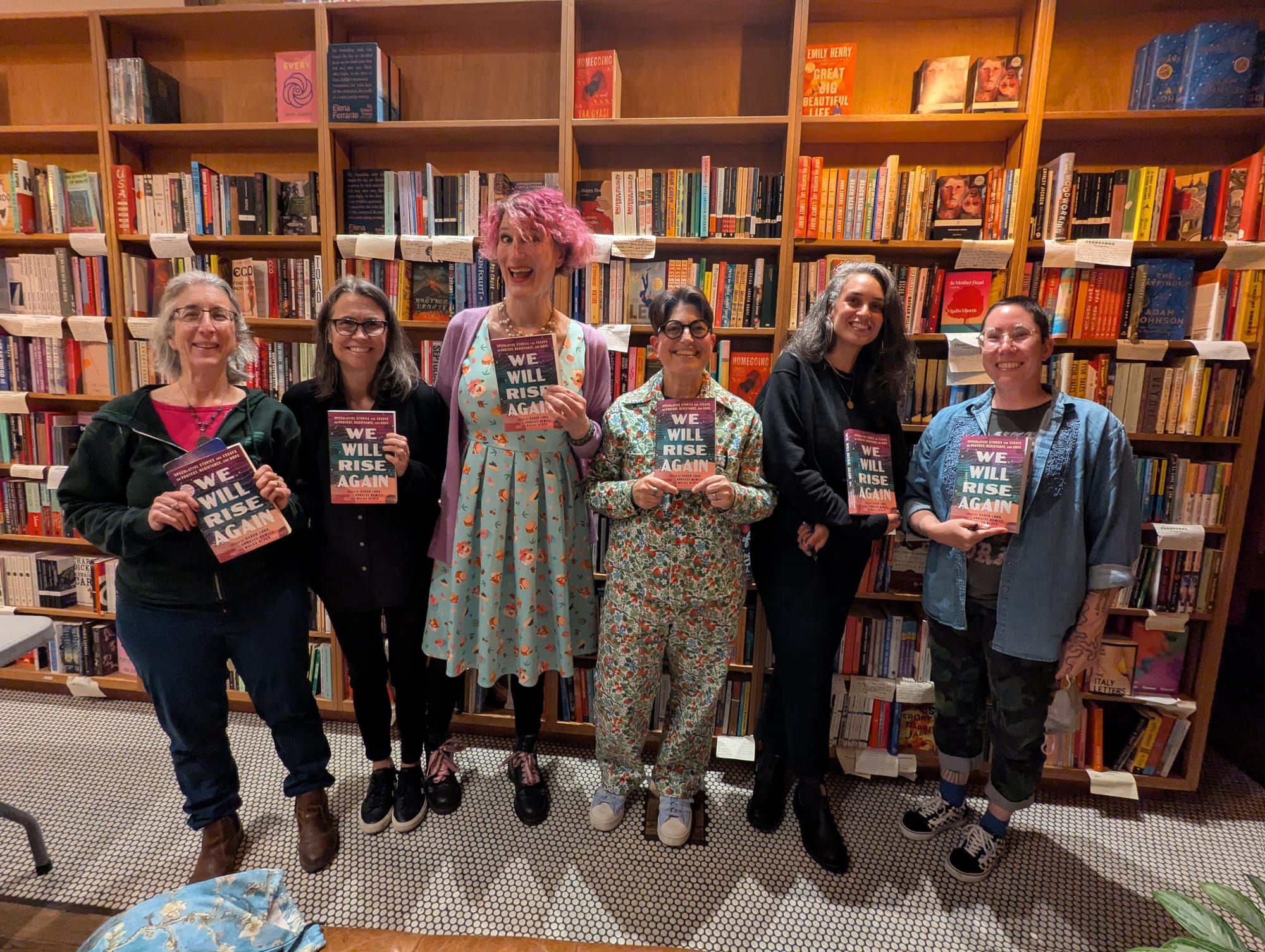 Six people all holding a copy of the same book (We Rise Again) standing in front of a bookstore wall of books and looking very excited.