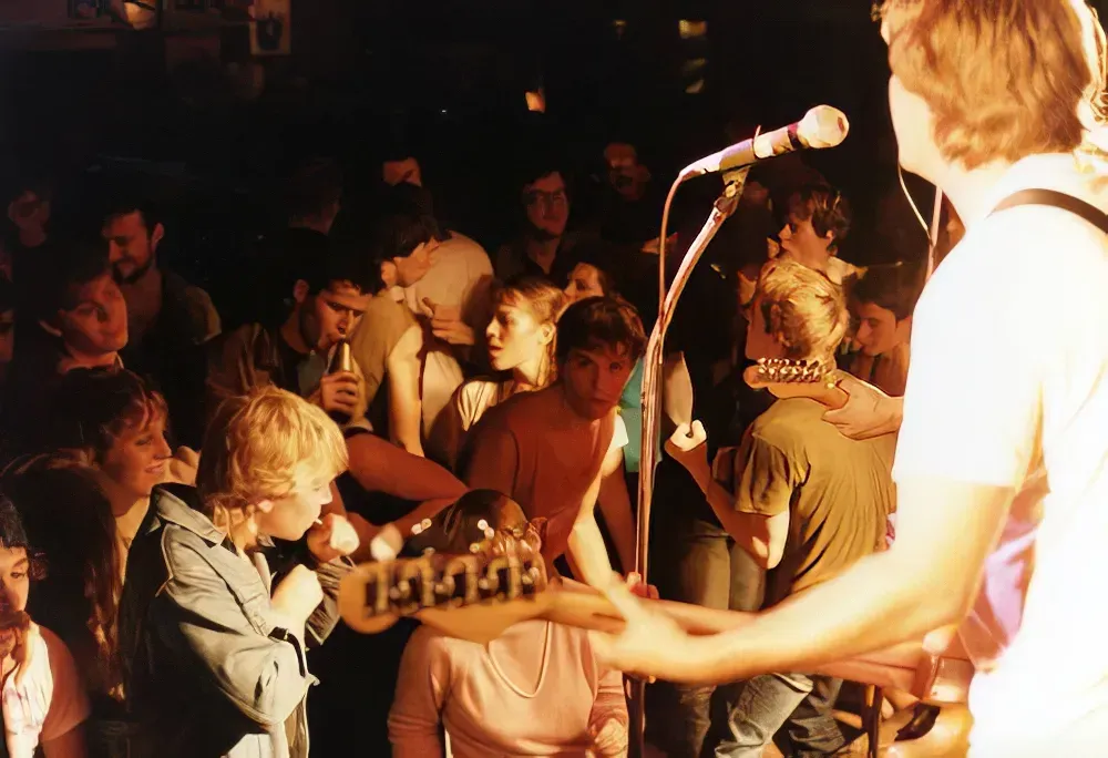 1980s punk show crowd packed in front of a small stage, musicians playing and audience members pressed close in a dim, chaotic room