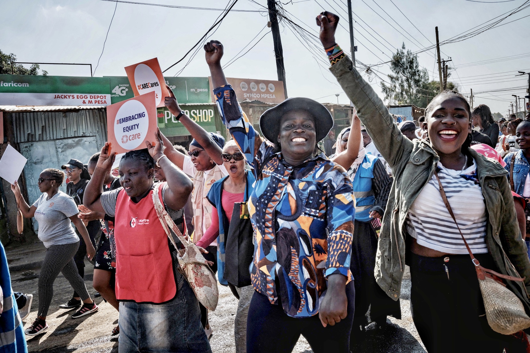Two women smile at the camera while holding their fists up. Besides them another person holds a paper that reads, "#Embracing Equity. CARE".