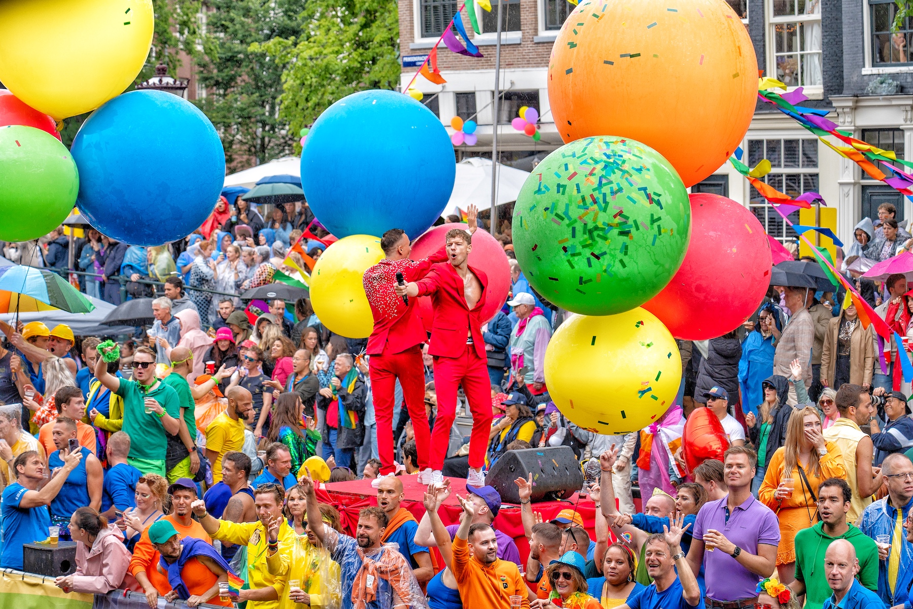 Two men dressed in red are on a stage performing. People in colorful clothes surround them. 