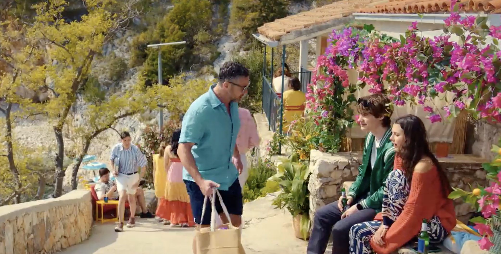 Joe (Warren Brown inspects Daniele Russo (Alessandro Bedetti) who his daughter Angelica (Beau Gadsdon) has befriended. They're sitting on a bench by a path near the beach with pink blooms in the background 