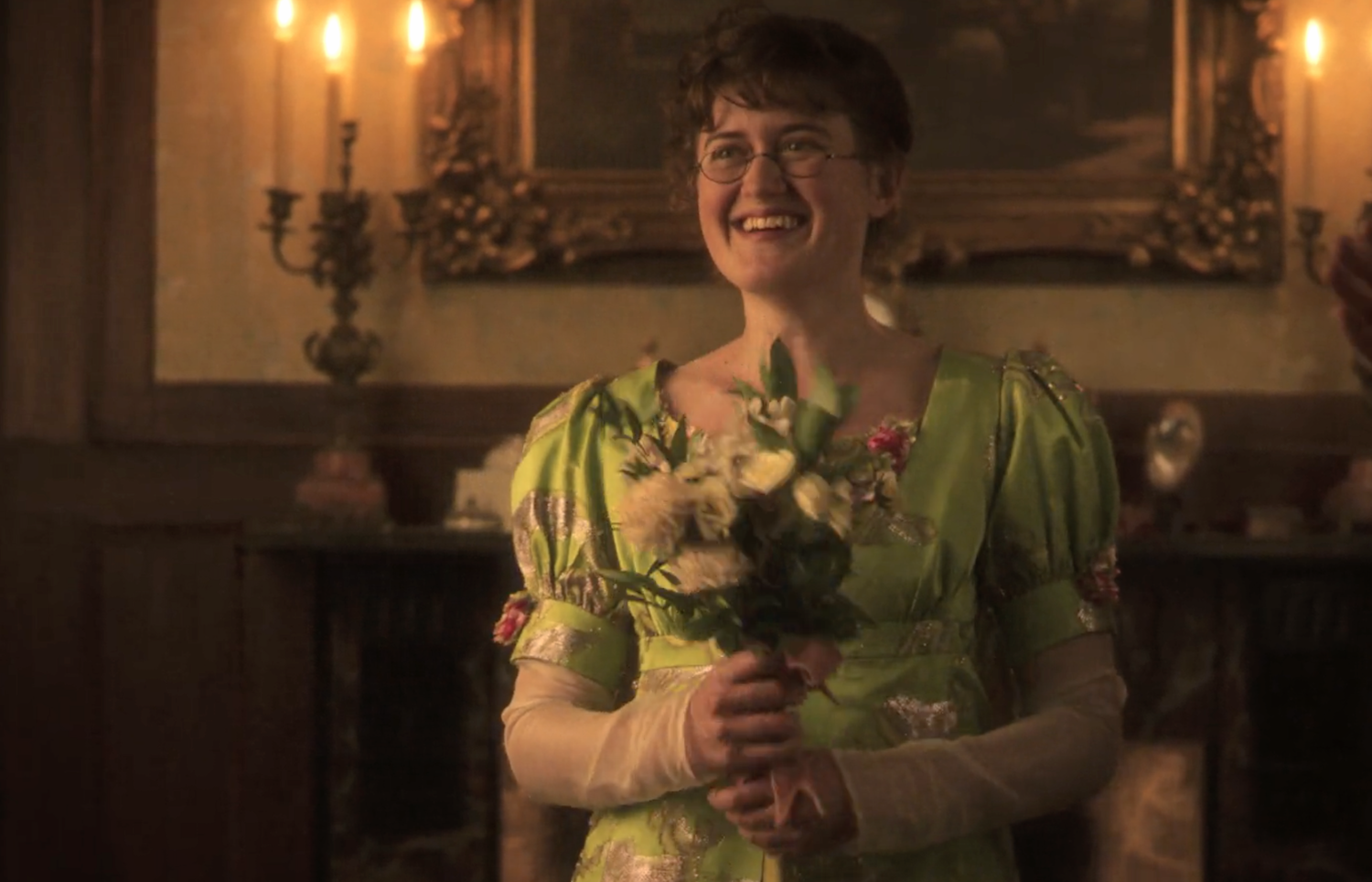 Mary Bennet (Ella Bruccoleri) in front of a fireplace and mirror, holds a bunch of flowers
