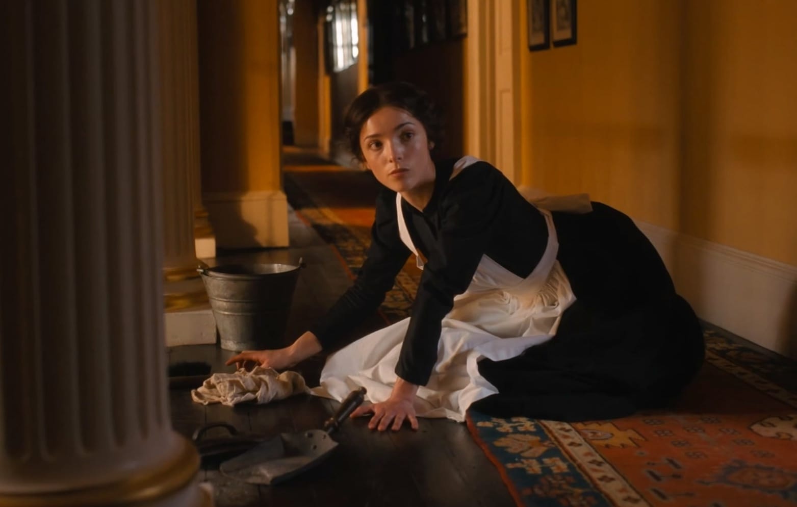 Emma (Jessica Reynolds) in her maid's uniform of black dress and white apron, scrubs the floor.