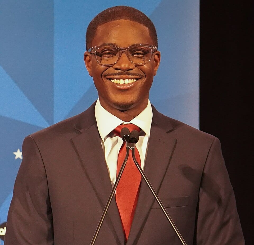 A picture of a smiling Black man in a suit, standing before two microphones. 