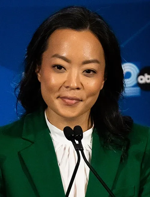 A picture of an Asian American woman in a green blouse with two microphones before her.