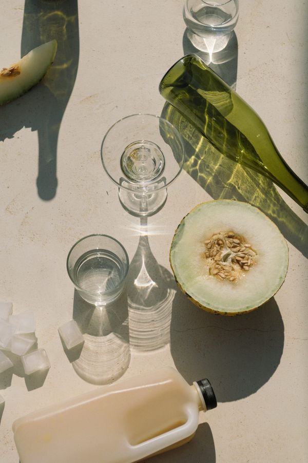 Photograph of some glasses, a plastic jug, a few ice cubes, and half of a melon on a white surface