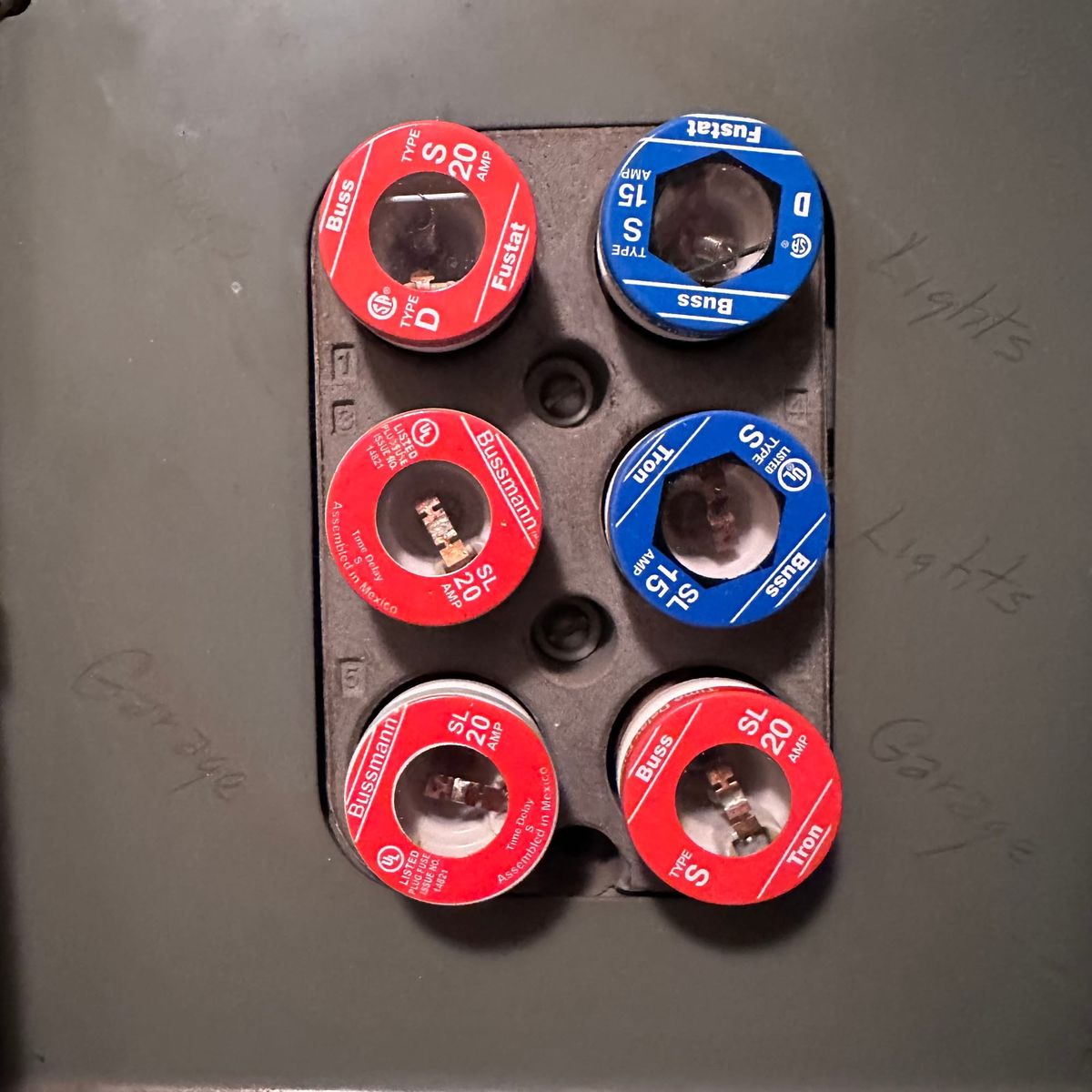 6 ancient screw-in fuses in an ancient fuse box. There are 4 red & two blue fuses. There's light pencil beside some saying garage or lights.