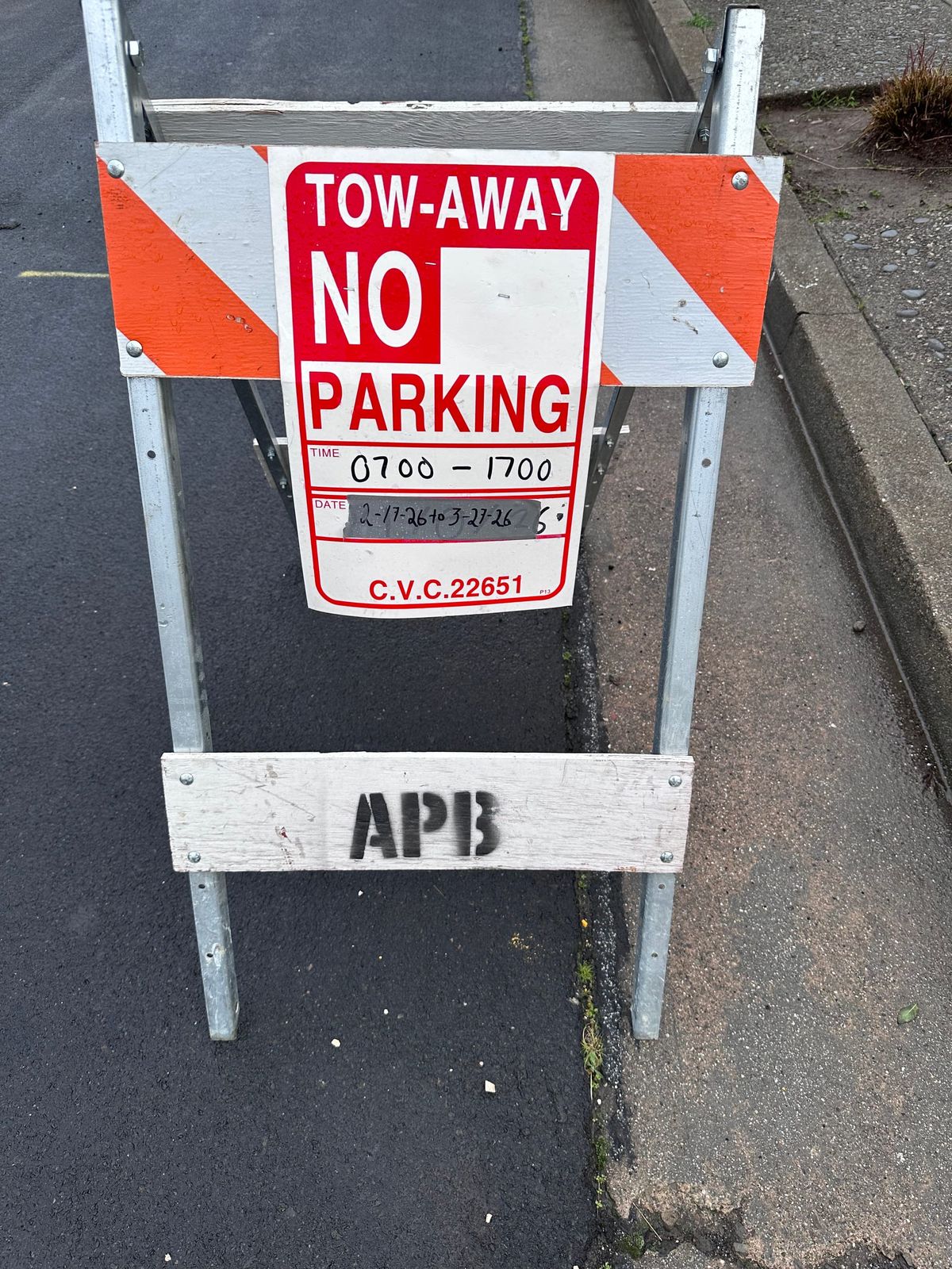 an a-frame in a wet street. APB is stencilled on the bottom crossbar & the top has a "tow away No Parking" sign. The time is 0700 - 1700 the date is duct-tape w/sharpie 2-17-26 to 3-27-26
