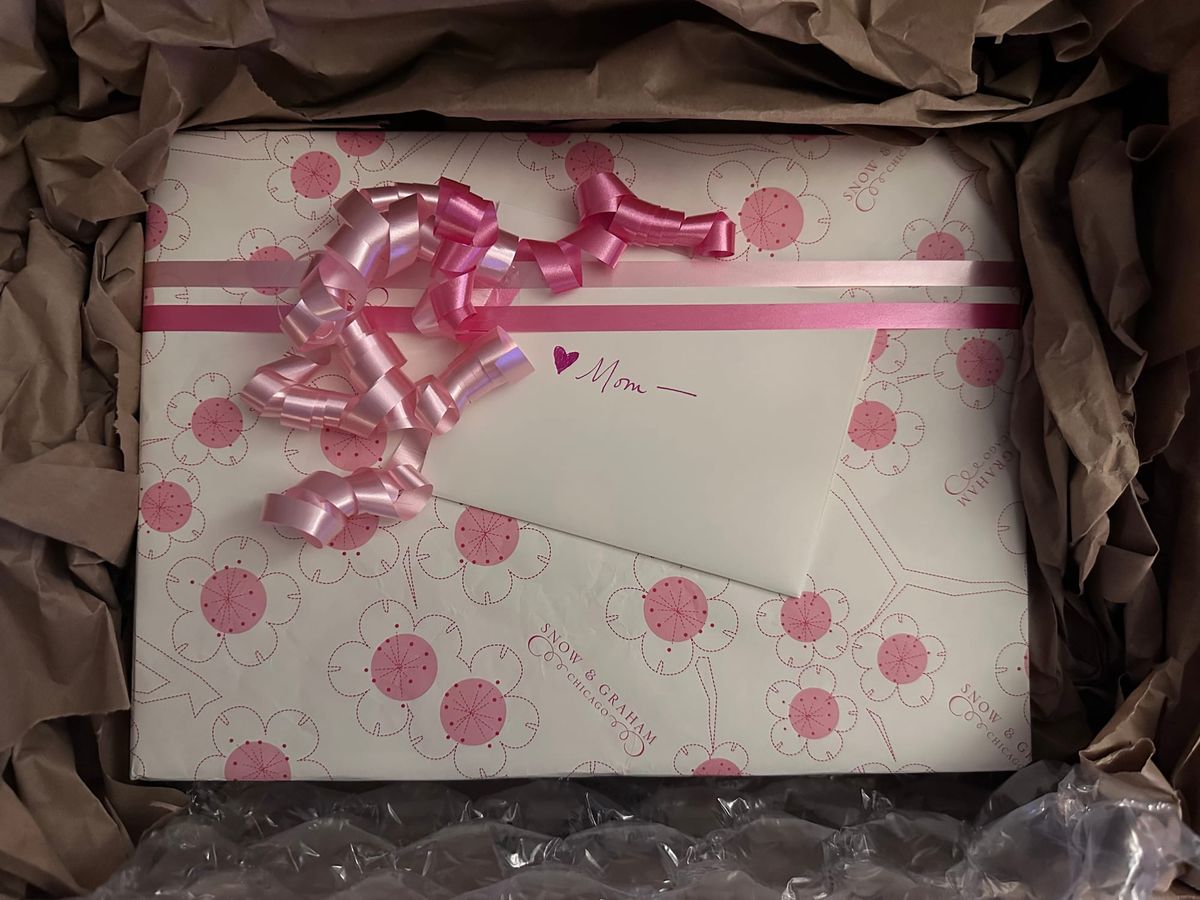 A wrapped box in white paper with pink and fuchsia flowers printed on it, curly ribbon, a card with handwritten "mom" on it, all nestled in packing paper.