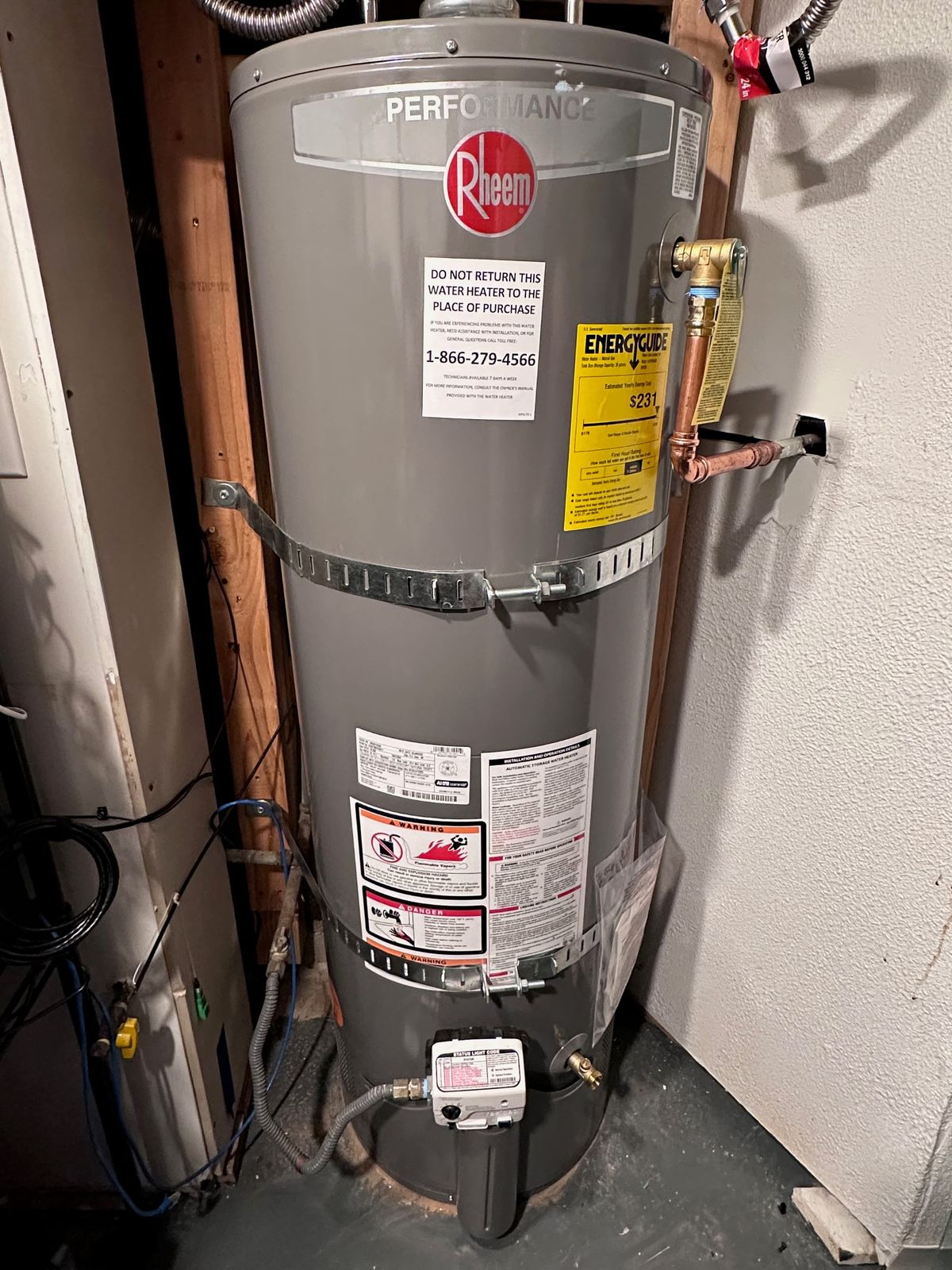A new shiny grey tank water heater with pipes, clamps, hoses all with tags and new stickers all over it, set into a basement corner. It says "performance rheem" in a shiny label at top.