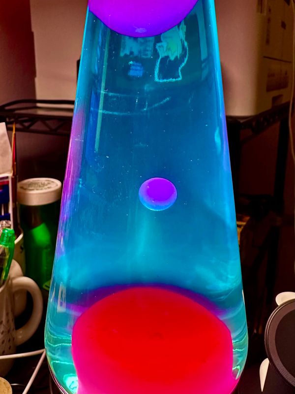 Closeup of a lava lamp with blue fluid and pink lava in mid bloop. You can see some desk cruft in the background around it. A laser printer on a stand is visible behind the fluid.