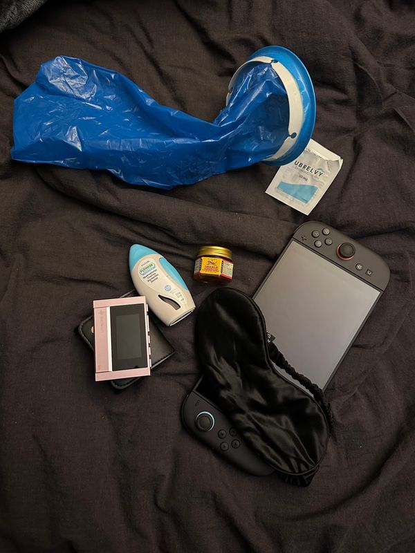 Various items on a black duvet: Nintendo Switch 2, eyemask, small pink media player, nose spray, tiger balm, barf bag, Ubrelvy packet.
