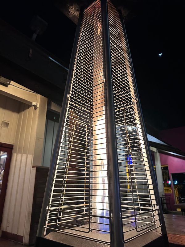 A pyramidal patio heater with a flame inside. There is a restaurant patio behind the heater & in the UR corner is the crescent moon in a dark sky.