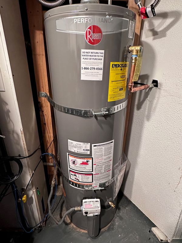 A new shiny grey tank water heater with pipes, clamps, hoses all with tags and new stickers all over it, set into a basement corner. It says "performance rheem" in a shiny label at top.