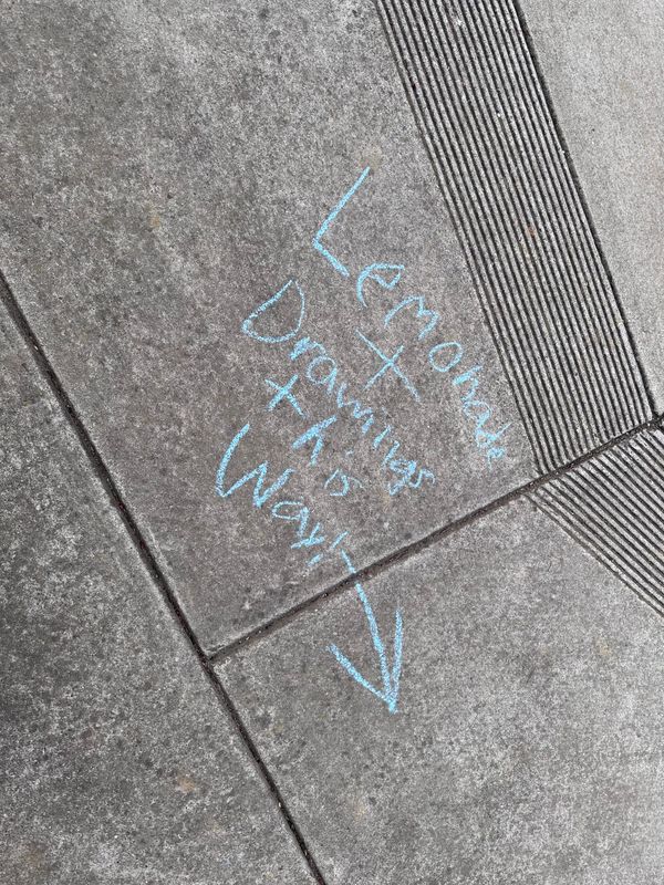 Portrait photo of a sidewalk with blue chalk on it that says "Lemonade + Drawings this way!" with an arrow pointing down.
