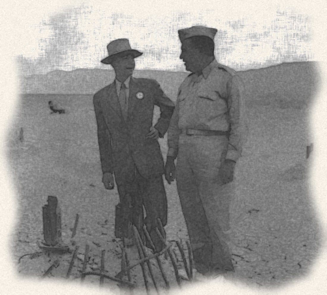 戴帽、穿西裝的歐本海默與穿軍服的格羅夫斯將軍，站在沙漠裡試爆塔殘留的鋼筋旁交談
