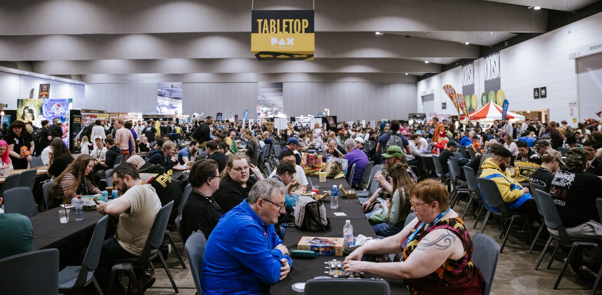 A large hall full of folks playing board games together. A sign hangs above: "Tabletop PAX"