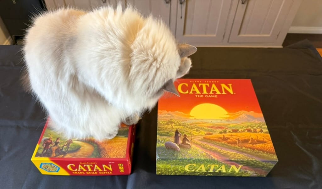 A white ragdoll cat has its faced turned down and away from the camera while sitting on the 5th edition Catan box, with the sixth edition Catan box more clearly visible to the right. There is a black tablecloth and in the background the lower part of a light brown cabinet.